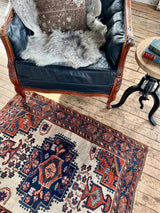 Antique Persian area rug with intricate patterns on a wooden floor, featuring a black leather chair with sheepskin.
