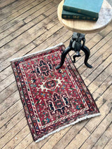 Small red Persian Karajah rugon a wooden floor with a small table and book in the background.