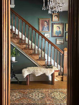 A classic wooden staircase with wood railings, set against an olive green wall adorned with framed portraits. A sheepskin rests on a wooden bench at the base of the stairs, with a vintage Persian runner rug on the floor