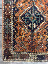 Corner of a traditional hand-knotted vintage Persian rug with a geometric pattern, featuring a prominent central diamond medallion in dark navy blue, teal, and cream, surrounded by an orange/rust field and a dark brown border.