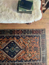 A hand-knotted vintage Persian rug with a geometric pattern, featuring a prominent central diamond medallion next to a bench with sheepskin and books