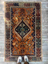 A traditional hand-knotted vintage Persian rug with a geometric pattern, featuring a prominent central diamond medallion in dark navy blue, teal, and cream, surrounded by an orange/rust field and a dark brown border.