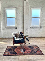 Vintage Persian Saveh area rug with dusty pink and blue medallions and a navy field on a wooden floor with a blue armchair and white brick walls.