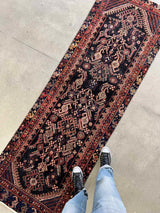 Person wearing black sneakers and light blue jeans standing on a vintage Persian Hamadan runner rug with brick red and peach herati motif and navy blue field