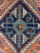 Close-up of a hand-knotted vintage Persian rug with a geometric pattern, featuring a prominent central diamond medallion in dark navy blue, teal, and cream, surrounded by an orange/rust field and a dark brown border.