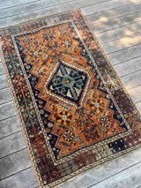 A traditional hand-knotted vintage Persian rug with a geometric pattern, featuring a prominent central diamond medallion in dark navy blue, teal, and cream, surrounded by an orange/rust field and a dark brown border.