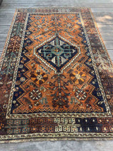 A hand-knotted vintage Persian rug with a geometric pattern, featuring a prominent central diamond medallion in dark navy blue, teal, and cream, surrounded by an orange/rust field and a dark brown border.