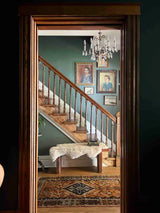 A dark wood doorway frames an interior view of a staircase, a green wall, framed portraits, a hanging chandelier, and a vintage Persian  accent  rug.